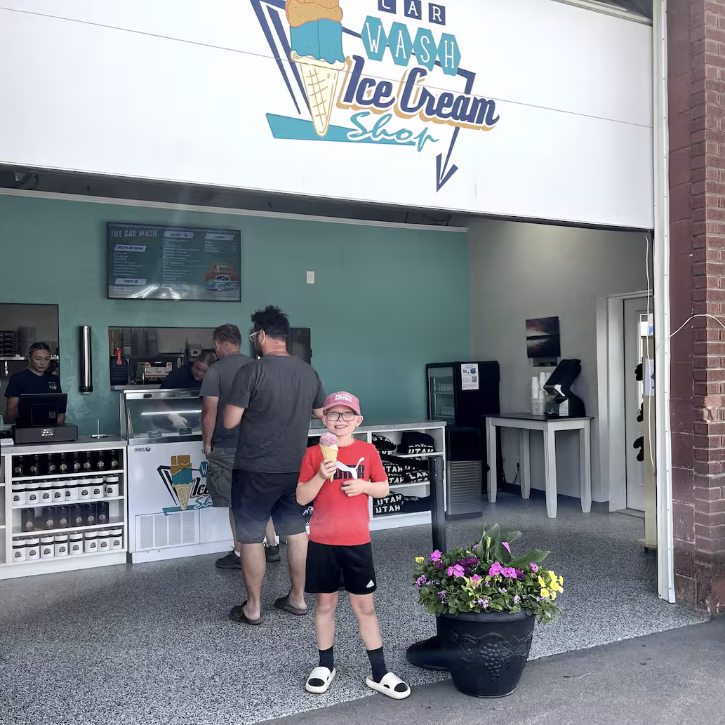 A young boy in a red shirt and cap holds an ice cream cup and smiles outside the “Car Wash Ice Cream Shop” at a nearby luxury glamping resort. People order inside, while a flower pot sits nearby, adding charm to the scene.
