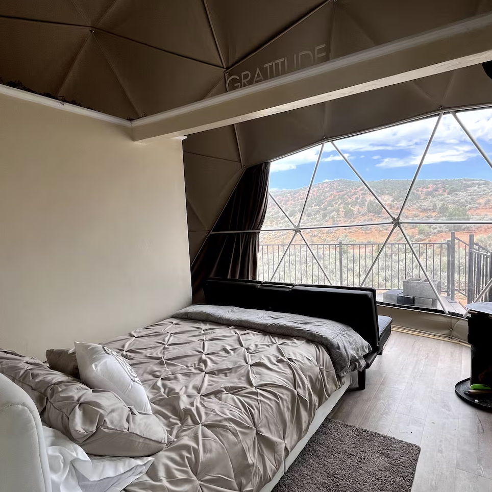 A cozy bed faces a large geometric window with sweeping views of mountains and a bright sky. This Glamping Resort features soft bedding, a dark sofa, and modern decor, creating a relaxing, scenic retreat in luxury glamping style.