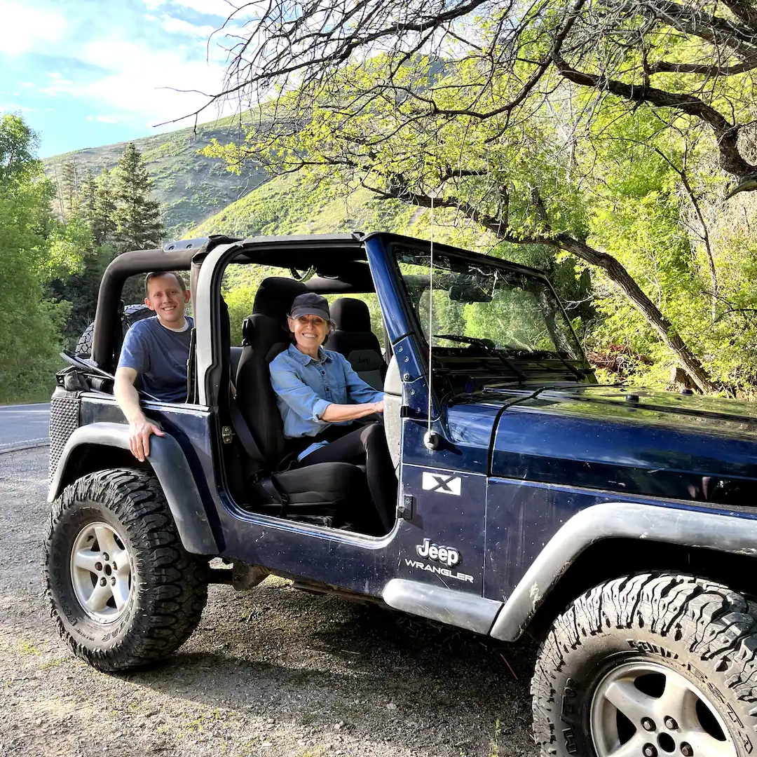 Two people smile in and next to a dark blue Jeep Wrangler parked on a scenic road near a luxury glamping resort, surrounded by green trees and hills with sunlight filtering through the branches above.