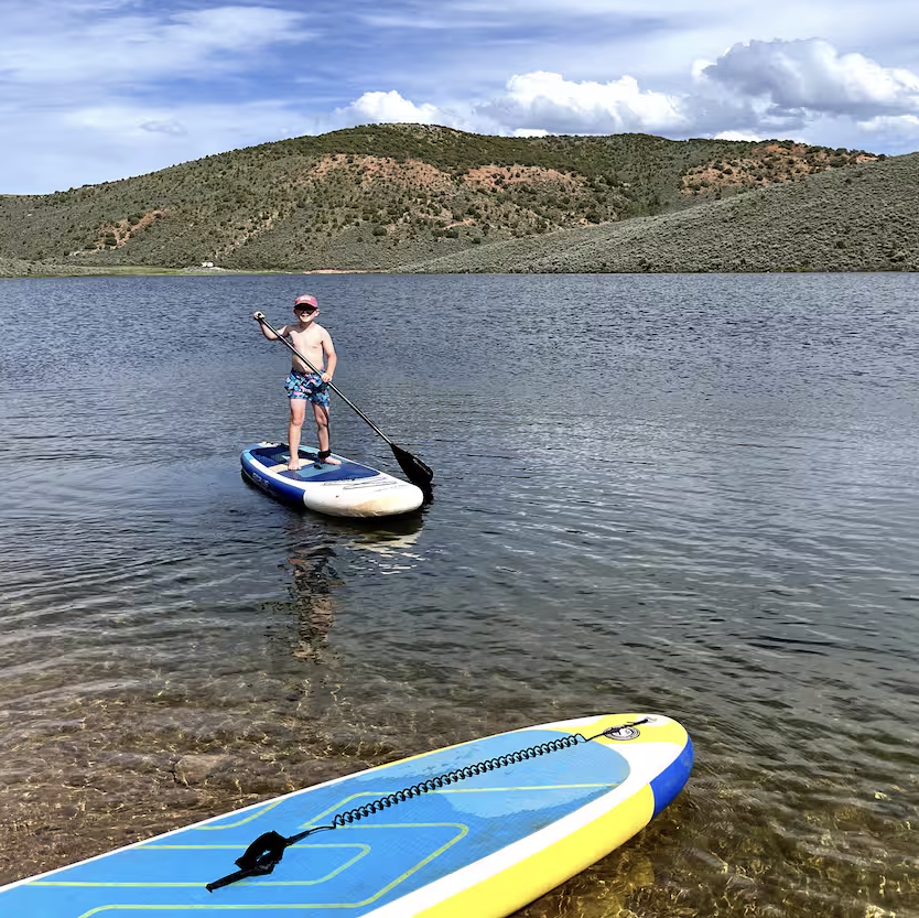 A child wearing a hat and swim trunks paddleboards on a calm lake near a rocky shore at a luxury glamping resort, while another paddleboard rests in the shallow water. Hills and clouds provide a scenic backdrop.