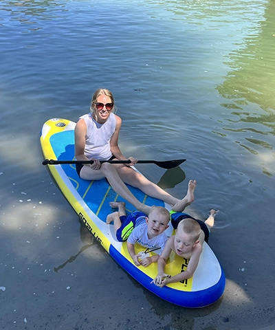 A woman wearing sunglasses sits on a paddleboard in shallow water at Bear Lake Utah, holding a paddle, with two young boys in swimwear lying at the front. Sunlight reflects off the water—a perfect day near a luxury glamping resort.