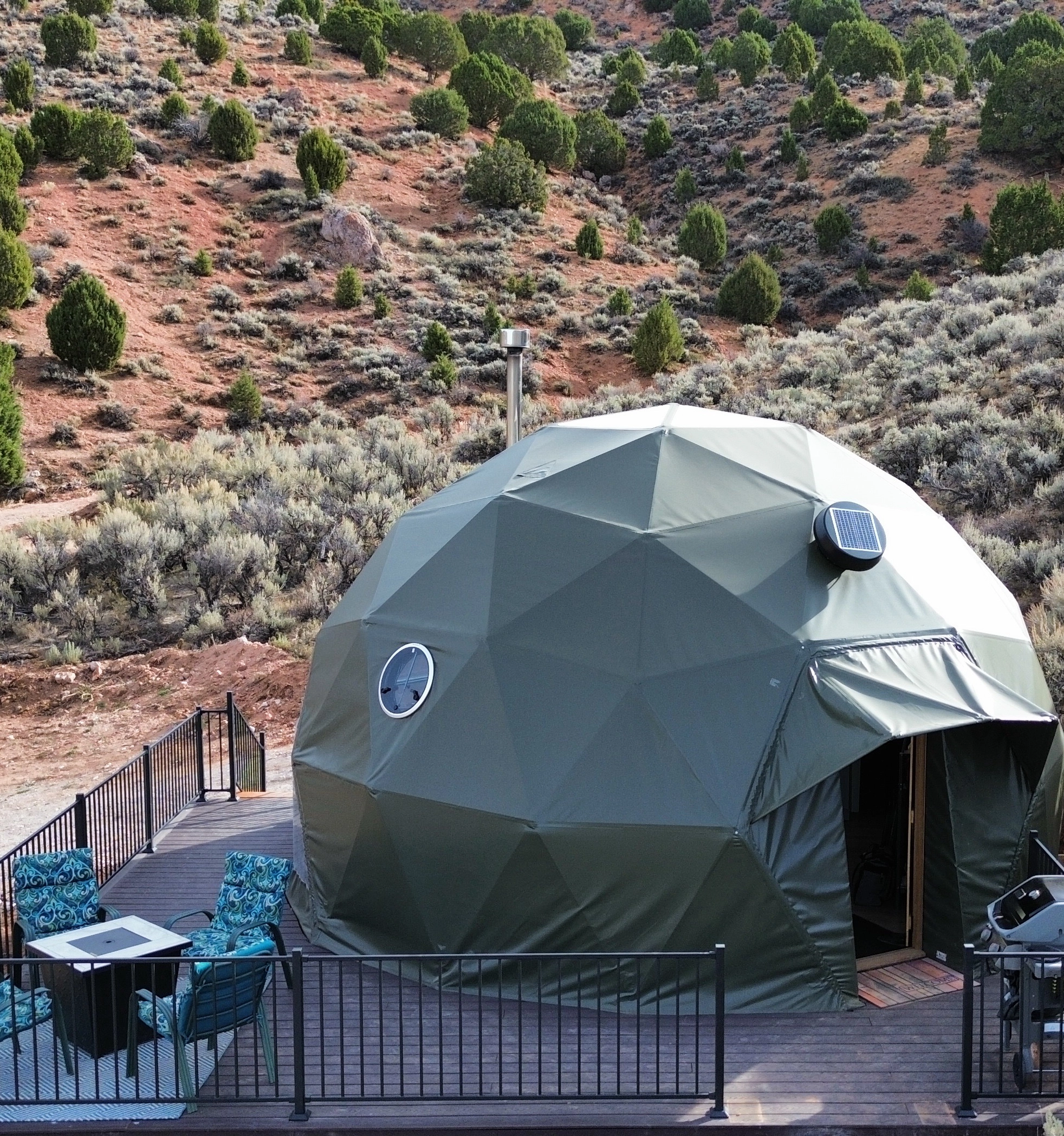 A green geodesic dome tent sits on a wooden deck with outdoor seating, surrounded by dry shrubs and small trees in a rocky, semi-arid landscape at a luxury glamping resort near Bear Lake Utah.