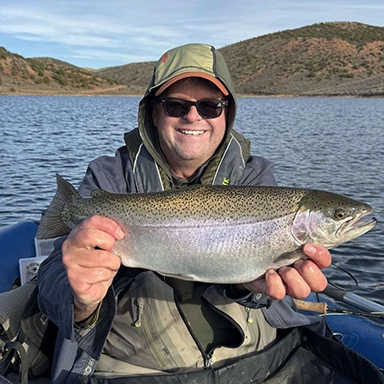 A smiling person wearing sunglasses and a hooded jacket holds a large fish on a boat at Bear Lake Utah, with the scenic lake and hills in the background under a partly cloudy sky.
