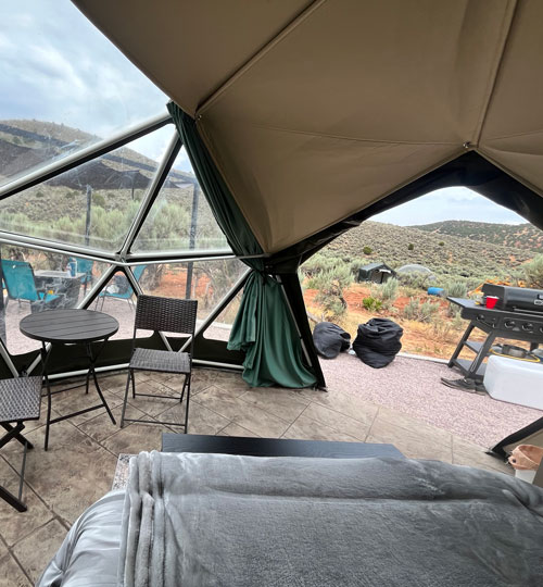 Interior view of a geodesic dome tent with a small table and chairs, a bed, and outdoor scenery of shrubs and mountains visible through large, clear windows. A grill and more chairs are outside on a gravel area.