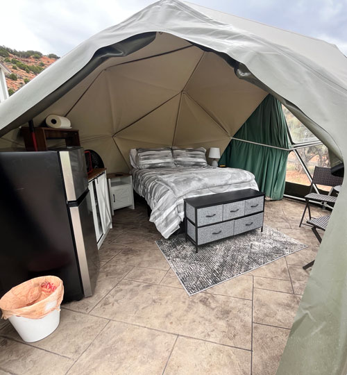A cozy glamping dome features a bed with gray bedding, a small dresser, a mini fridge, nightstand, rug, and chairs on a tiled floor. The dome has canvas walls with a window showing outdoor scenery.