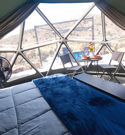 A cozy geodesic dome room with a bed, a small table set for two, and a view of a pool and dry, rocky hills through large geometric windows.