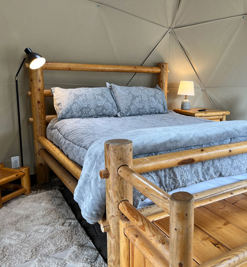 A cozy bedroom with a wooden log bed frame, light blue patterned bedding, two bedside tables with lamps, and a textured rug, set against beige geometric walls.